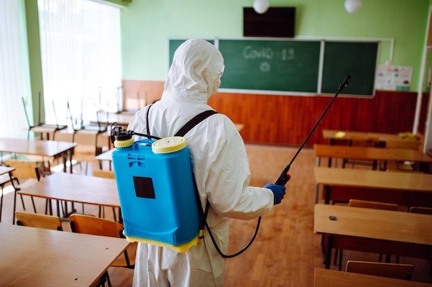 Vista posteriore di un operatore sanitario professionista che disinfetta l'aula prima dell'inizio dell'anno accademico. Un uomo che indossa una tuta protettiva pulisce l'auditorium dal coronavirus Covid-19. Assistenza sanitaria.