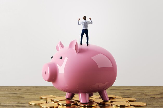 Vista posteriore di un piccolo uomo d'affari felice che celebra il successo in piedi su un porcellino rosa e monete d'oro sullo sfondo di un muro di cemento bianco raggiungendo gli obiettivi di investimento