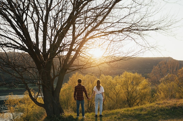 Vista posteriore di un ragazzo e una ragazza stanno insieme e si tengono per mano sotto un grande vecchio albero contro il muro di una foresta, collina e fiume al tramonto.