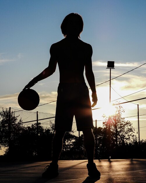 Vista posteriore di un uomo a silhouette che gioca a basket in campo durante il tramonto