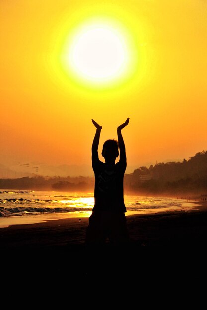 Vista posteriore di un uomo a silhouette con le braccia alzate sulla spiaggia durante il tramonto