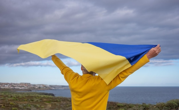 Vista posteriore di un uomo anziano dai capelli bianchi in piedi all'aperto sventolando la bandiera ucraina guardando il mare sentendo la libertà Nessuna guerra vogliamo la pace