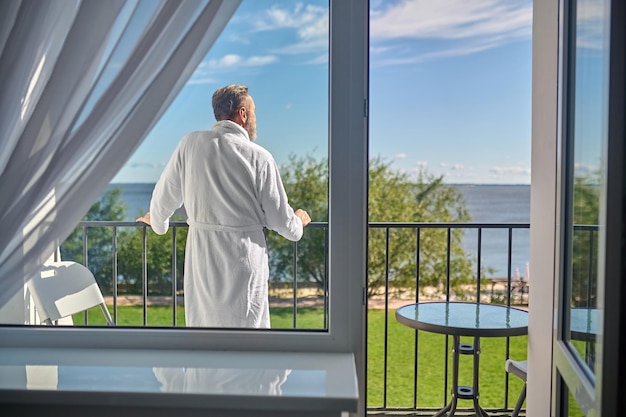 Vista posteriore di un uomo con l'accappatoio di spugna bianco appoggiato alla ringhiera del balcone