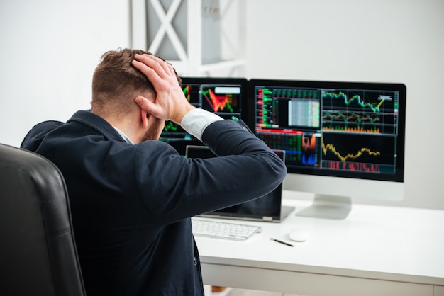 Vista posteriore di un uomo d'affari scioccato e stressato seduto e tenendo la testa per mano sul posto di lavoro in ufficio