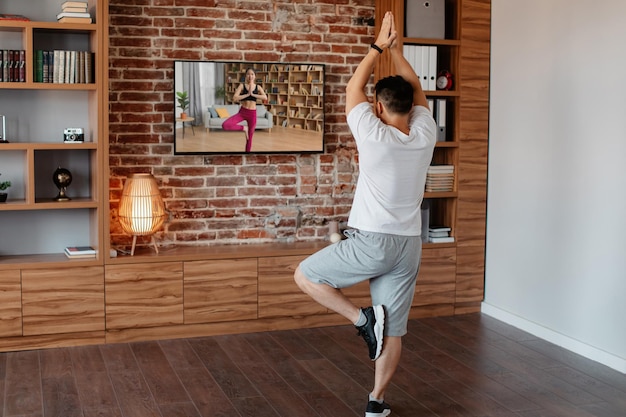 Vista posteriore di un uomo maturo che fa yoga mentre si guarda l'allenamento video in TV in piedi nella posa dell'albero con le braccia alzate