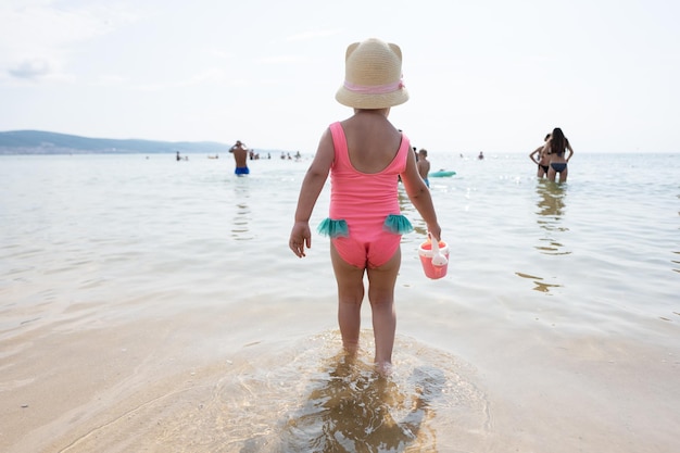 Vista posteriore di una bambina in costume da bagno rosa e un cappello di paglia in piedi nell'acqua sulla spiaggia