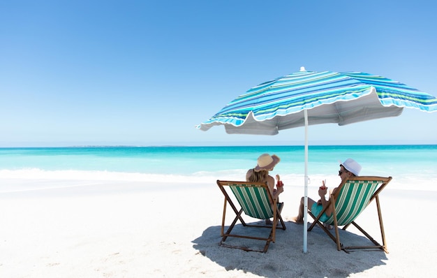 Vista posteriore di una coppia caucasica seduta su sedie a sdraio sotto l'ombrello, sulla spiaggia con il cielo blu e il mare sullo sfondo, sorridendo, guardandosi e facendo un brindisi