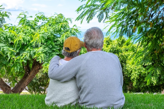 Vista posteriore di una coppia senior caucasica rilassata seduta nel prato nel parco pubblico che si gode le vacanze o il pensionamento nel tempo libero