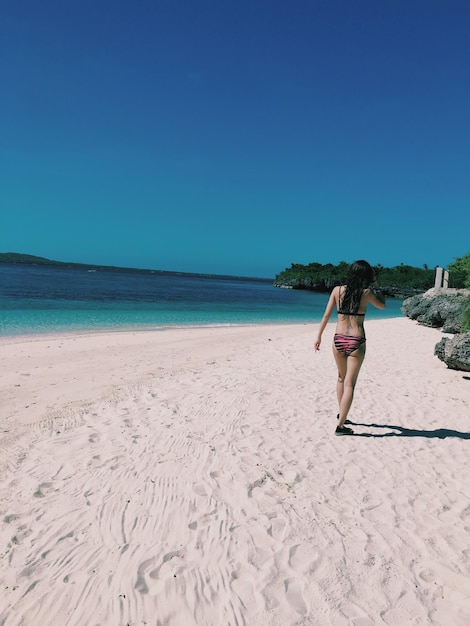 Vista posteriore di una donna che indossa un bikini che cammina sulla spiaggia contro un cielo blu limpido