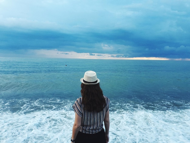 Vista posteriore di una donna che indossa un cappello che guarda il mare contro un cielo nuvoloso