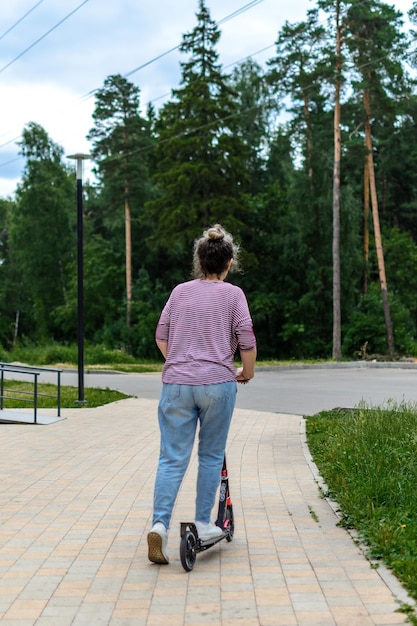 Vista posteriore di una donna che si rilassa in un parco vicino alla foresta, in sella a uno scooter Kick, spazio per la copia.