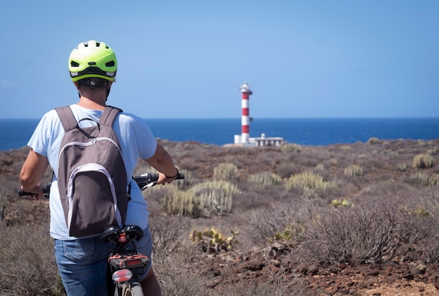 Vista posteriore di una donna con casco giallo godendo di andare in bicicletta vicino a un faro in vacanze al mare - paesaggio arido e orizzonte sull'acqua in una giornata di sole a Tenerife, Isole Canarie