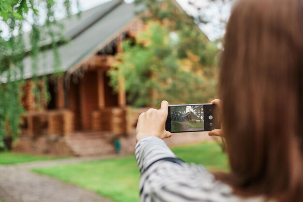 Vista posteriore di una donna dai capelli castani che fotografa la casa di legno in parte visitando nuovi posti in giro per la città con video di riprese per le vacanze per il suo blog di viaggio