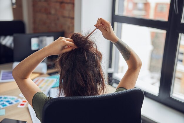 Vista posteriore di una donna impegnata al lavoro che fa una crocchia per capelli usando mani femminili con bastoncini di legno e marrone disordinato