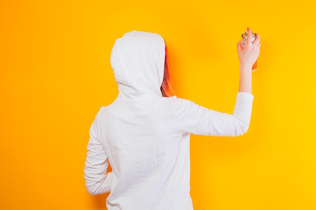 Vista posteriore di una donna in piedi su uno sfondo giallo con spray La giovane donna vestita con una felpa con cappuccio bianca disegna usando spray Il concetto di cultura giovanile