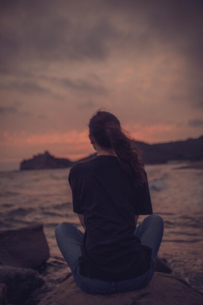Vista posteriore di una donna seduta su una roccia sulla spiaggia durante il tramonto