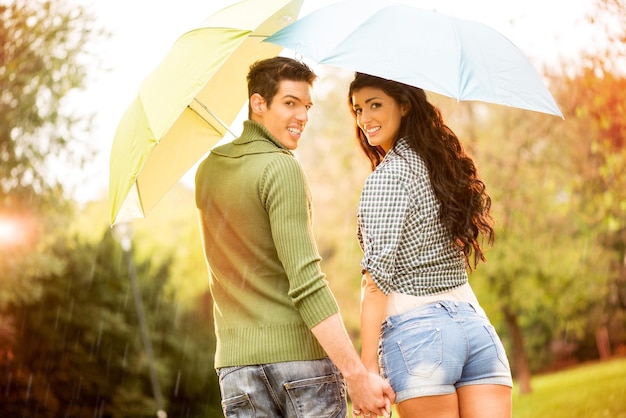 Vista posteriore di una giovane coppia innamorata degli ombrelli mentre cammina sotto la pioggia attraverso il parco tenendosi per mano e guardando la fotocamera con un sorriso.