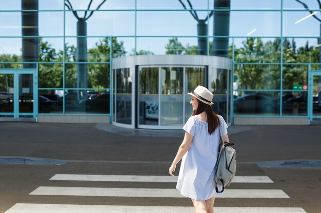 Vista posteriore di una giovane donna turistica sorridente con cappello con zaino in piedi sul passaggio pedonale all'aeroporto internazionale