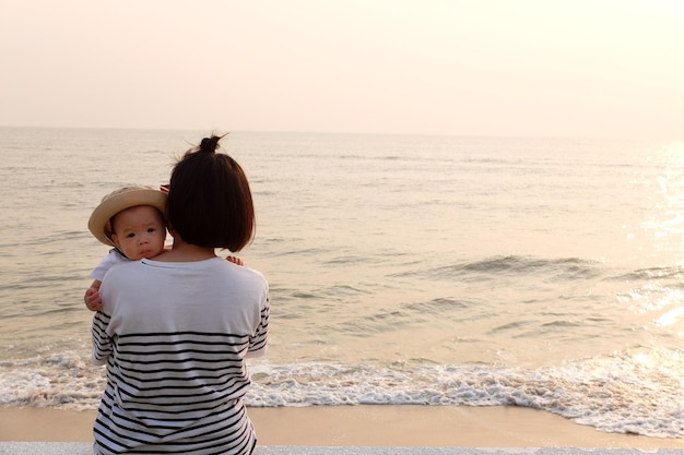 Vista posteriore di una madre che porta un bambino sulla spiaggia contro il cielo durante il tramonto