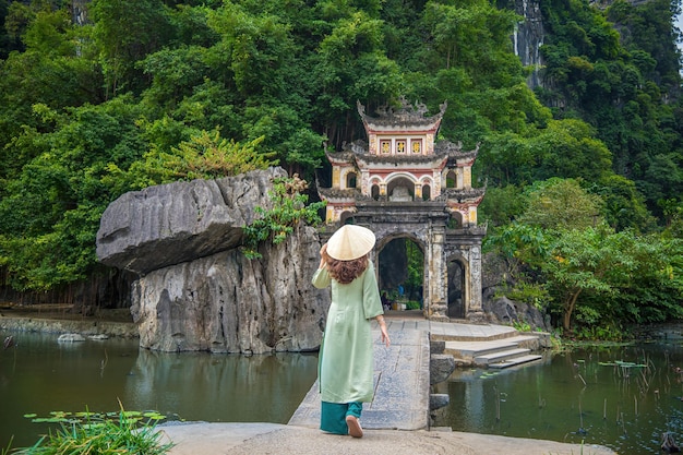Vista posteriore di una ragazza che indossa Ao Dai di fronte a un paesaggio di parco all'aperto con lago e ponte di pietra Entrata all'antico complesso della pagoda di Bich Dong destinazione di viaggio di Ninh Binh Vietnam