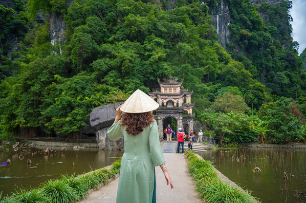 Vista posteriore di una ragazza che indossa Ao Dai di fronte a un paesaggio di parco all'aperto con lago e ponte di pietra Entrata all'antico complesso della pagoda di Bich Dong destinazione di viaggio di Ninh Binh Vietnam
