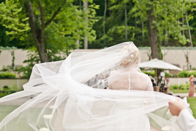 Vista posteriore di una sposa in un abito di pizzo con un velo che fluttua nel vento viene inviato al luogo della cerimonia di matrimonio