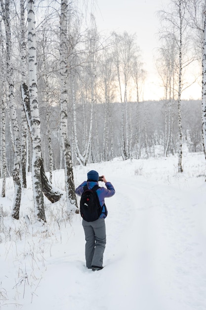 Vista posteriore di una studentessa in una calda giacca invernale e cappello che fotografa una bella foresta di betulle innevata