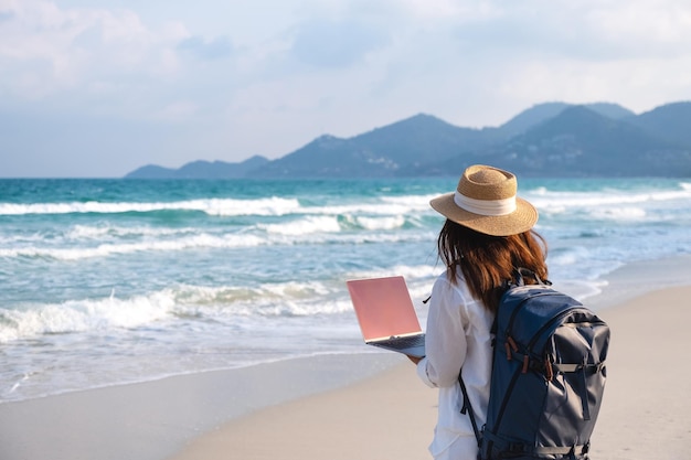 Vista posteriore di una viaggiatrice che usa e lavora al computer portatile mentre cammina sulla spiaggia