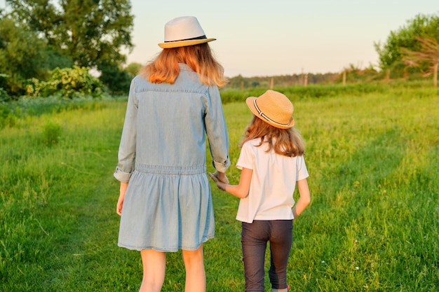 Vista posteriore, due bambine vanno tenendosi per mano in un prato verde estivo.