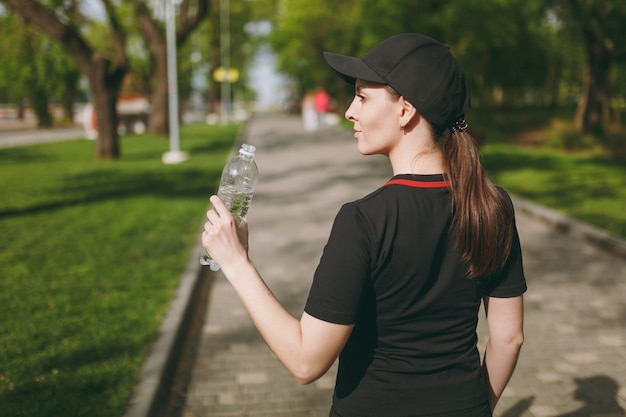 Vista posteriore Giovane bella donna bruna atletica in uniforme nera e berretto con bottiglia, con acqua durante l'allenamento prima di correre in piedi nel parco cittadino all'aperto