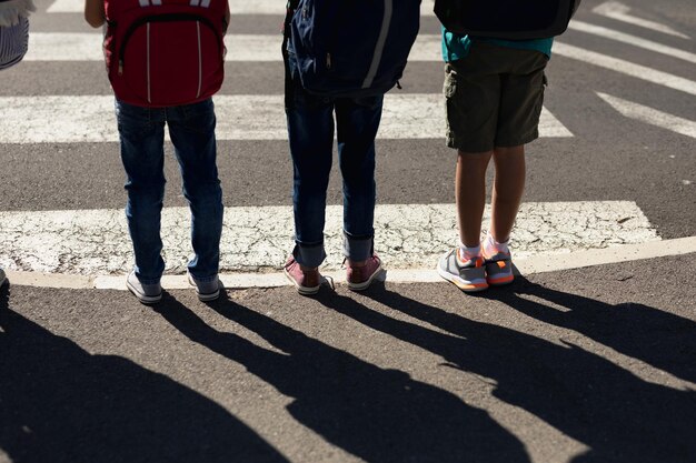 Vista posteriore in sezione bassa di un gruppo multietnico di tre scolari in attesa di attraversare la strada su un passaggio pedonale per andare a scuola. I bambini imparano la sicurezza stradale
