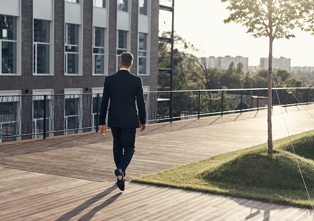 Vista posteriore integrale dell'uomo in abito completo che cammina vicino all'edificio per uffici all'aperto