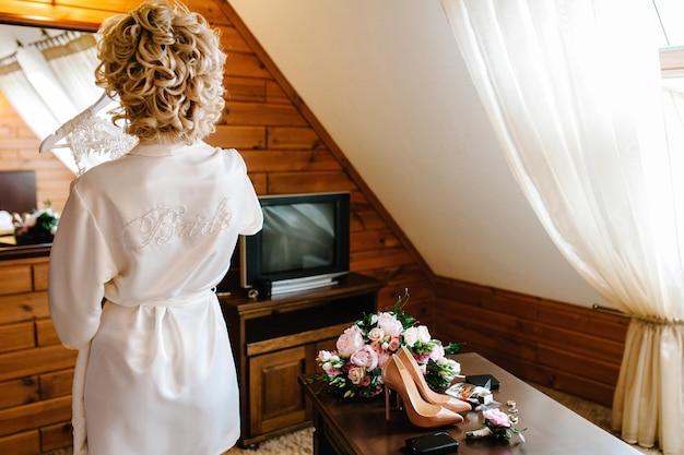 Vista posteriore la sposa tiene un abito da sposa nella stanza vicino allo specchio Ragazza elegante in stile in abito con pizzo Preparazioni mattutine per le nozze
