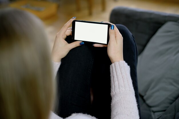 Vista posteriore ravvicinata di una mano di donna che tiene e utilizzando smart phone con schermo vuoto vuoto bianco seduto su un letto a casa. Foto di alta qualità