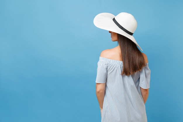 Vista posteriore ritratto di giovane donna di moda elegante in cappello da sole a tesa larga estate bianco, vestito messo le mani sulla testa, guardando da parte sullo spazio copia isolato su sfondo blu pastello. Concetto di stile di vita.