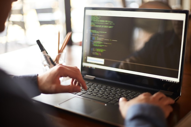 Vista posteriore sulla spalla di un programmatore sviluppatore con un portatile Codice del programma e dati dello script sullo schermo Giovane freelance con gli occhiali che lavora a un progetto in un caffè