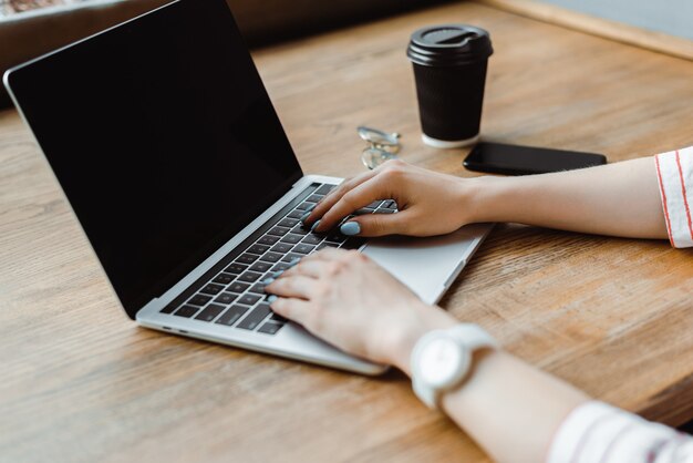 Vista potata della donna che utilizza computer portatile con lo schermo in bianco vicino allo smartphone e al caffè per andare alla tavola di legno nel caffè