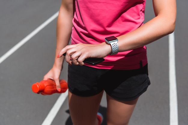 Vista potata della sportiva che esamina smartwatch mentre tiene la bottiglia di sport sulla pista corrente