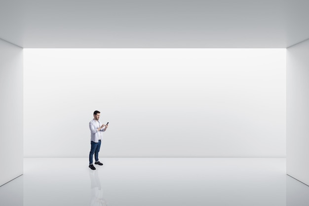 Vista prospettica sul giovane che utilizza lo smartphone in un'area vuota e spaziosa con sfondo bianco dello spazio della parete per il mock up della presentazione del prodotto o dell'auto
