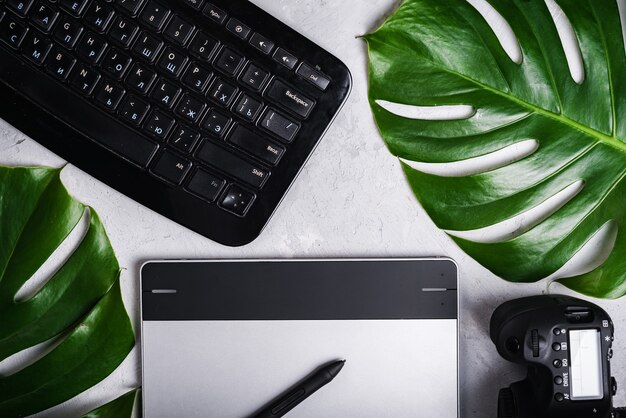 Vista ravvicinata del fotografo del posto di lavoro del designer grafico. Tablet, stilo, fotocamera, tastiera con lettere russe e inglesi, foglia verde monstera.