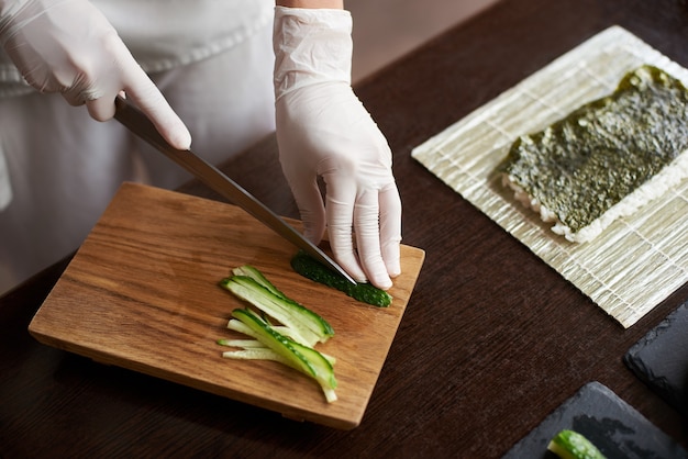 Vista ravvicinata del processo di preparazione di deliziosi sushi di laminazione nel ristorante. Mani femminili in guanti monouso per affettare il cetriolo sulla tavola di legno.