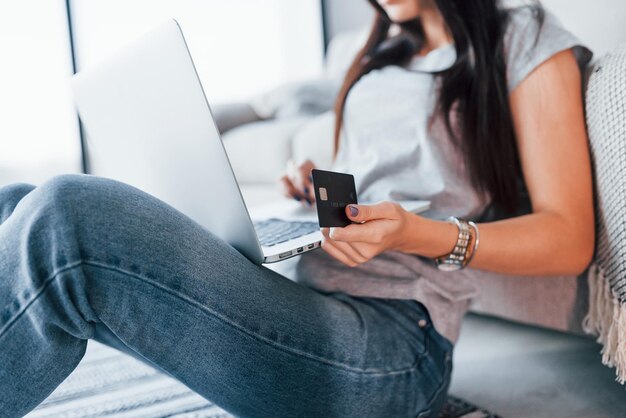 Vista ravvicinata della donna in abiti casual che è seduta a casa da sola con laptop e carta di credito.