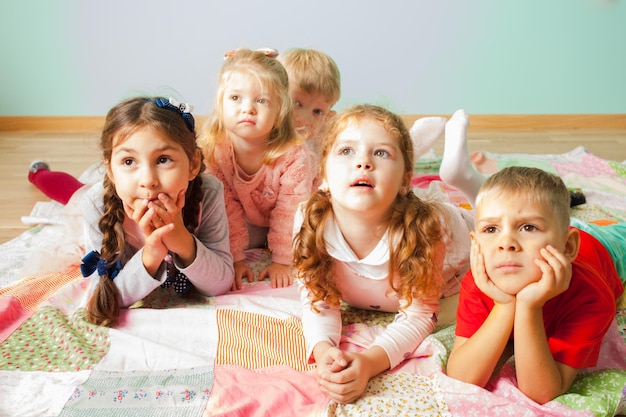 Vista ravvicinata di cinque adorabili bambini sdraiati su un pavimento su una copertina morbida che guardano con curiosità qualcosa dietro la telecamera. Gruppo di bambini in età prescolare che guardano cartoni animati in TV