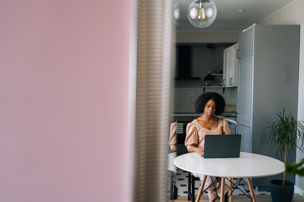 Vista remota di una giovane donna afroamericana seria che lavora o studia a distanza dall'ufficio domestico utilizzando l'ubicazione del laptop alla scrivania in cucina