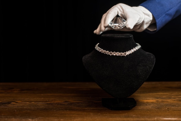 Vista ritagliata del perito di gioielli che tiene il diamante vicino alla collana sul supporto della collana sul tavolo di legno