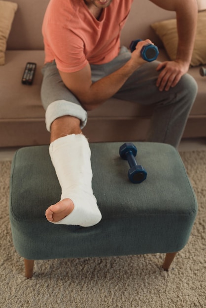 Vista ritagliata dell'uomo con la gamba rotta sul pouf che si allena con il manubrio sul divano