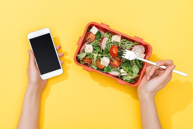 Vista ritagliata della donna con forchetta e pranzo al sacco con insalata utilizzando lo smartphone