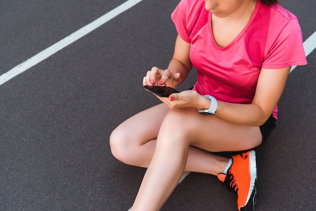 vista ritagliata della donna in smartwatch utilizzando smartphone sulla pista di atletica