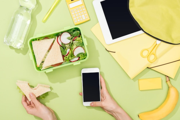 Vista ritagliata delle mani della donna con sandwich e smartphone vicino al pranzo al sacco con zaino alimentare digitale