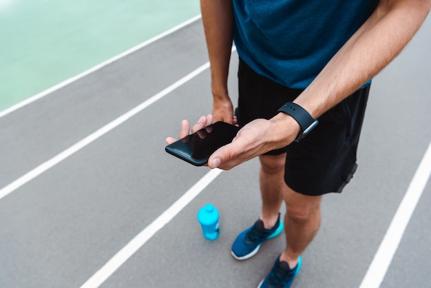 vista ritagliata di giovane sportivo che tiene smartphone con lo schermo in bianco sulla pista da corsa con bottiglia sport blu